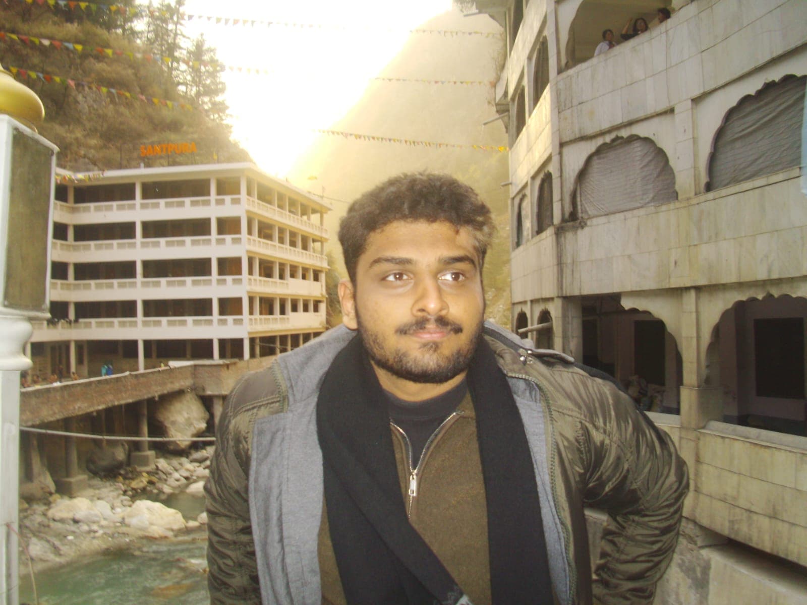 Rishi standing on a bridge in a mountain valley, wearing a jacket and scarf